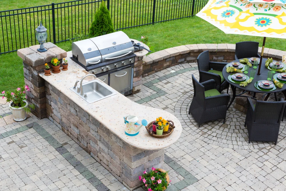 Outdoor patio with a stone kitchen featuring a grill and sink, surrounded by potted plants. Nearby, a round dining table with six wicker chairs sits under a colorful umbrella on a paved area with green grass outside the fence.