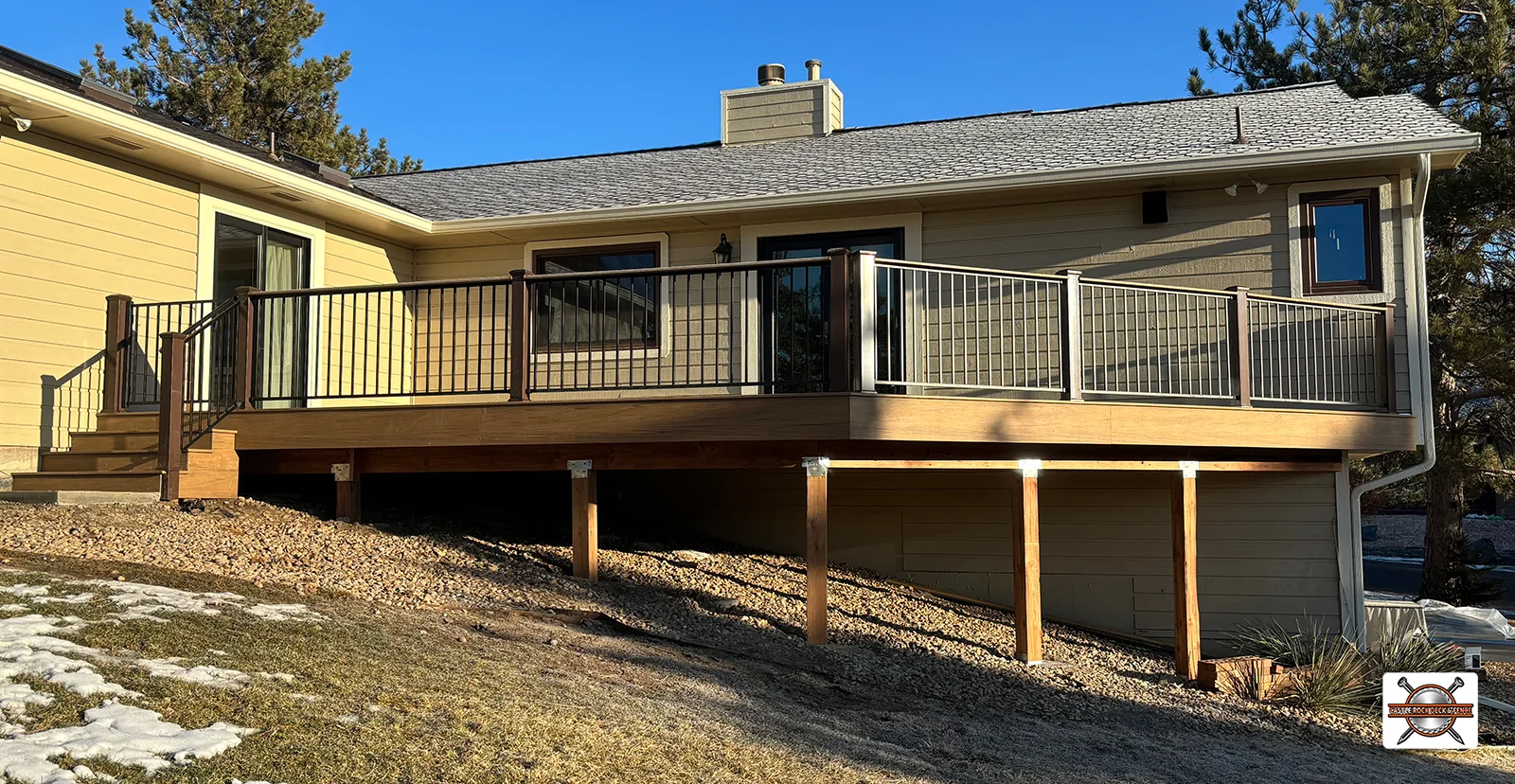 A wooden deck with metal railings is attached to a beige house, elevated on wooden posts over a sloped yard with some grass and rocks. Stairs lead from the deck to the ground.