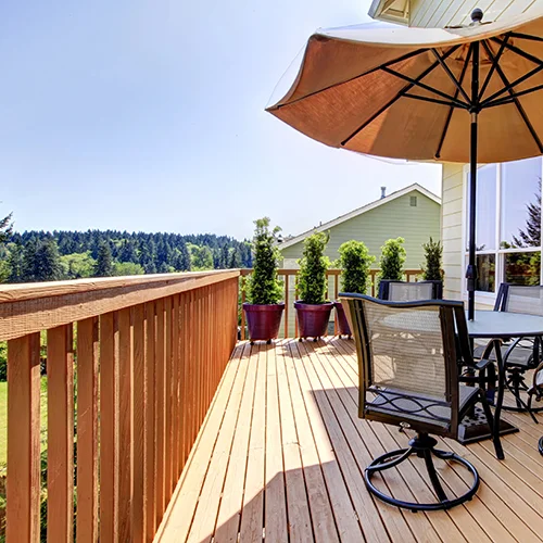 A sunny outdoor wooden deck with a round table, chairs, large umbrella, and potted plants. Trees and a neighboring house are visible in the background under a clear blue sky.