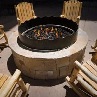 Four wooden rocking chairs arranged around a circular stone fire pit with a metal rim, containing burning flames and black stones, set on a stone patio.
