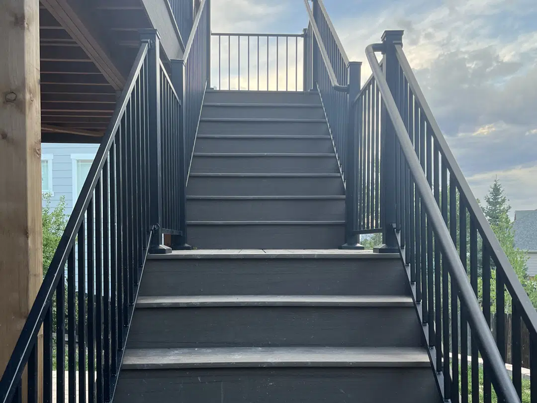A straight staircase with dark steps and black metal railings leads up to a landing outdoors, with greenery and a cloudy sky visible in the background.