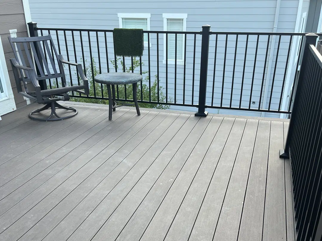 A gray patio with a black metal railing features a black rocking chair and a small round table. The deck overlooks a house with light blue siding and white-trimmed windows.