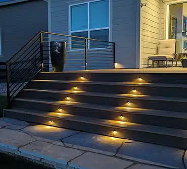 Modern outdoor steps with built-in warm yellow LED lights, leading up to a porch with chairs and a large black planter, in front of a gray house at dusk.