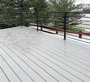 A clean, light gray composite deck with black metal railing, set outdoors on a slightly snowy day. Trees and a grassy area with patches of snow are visible in the background.