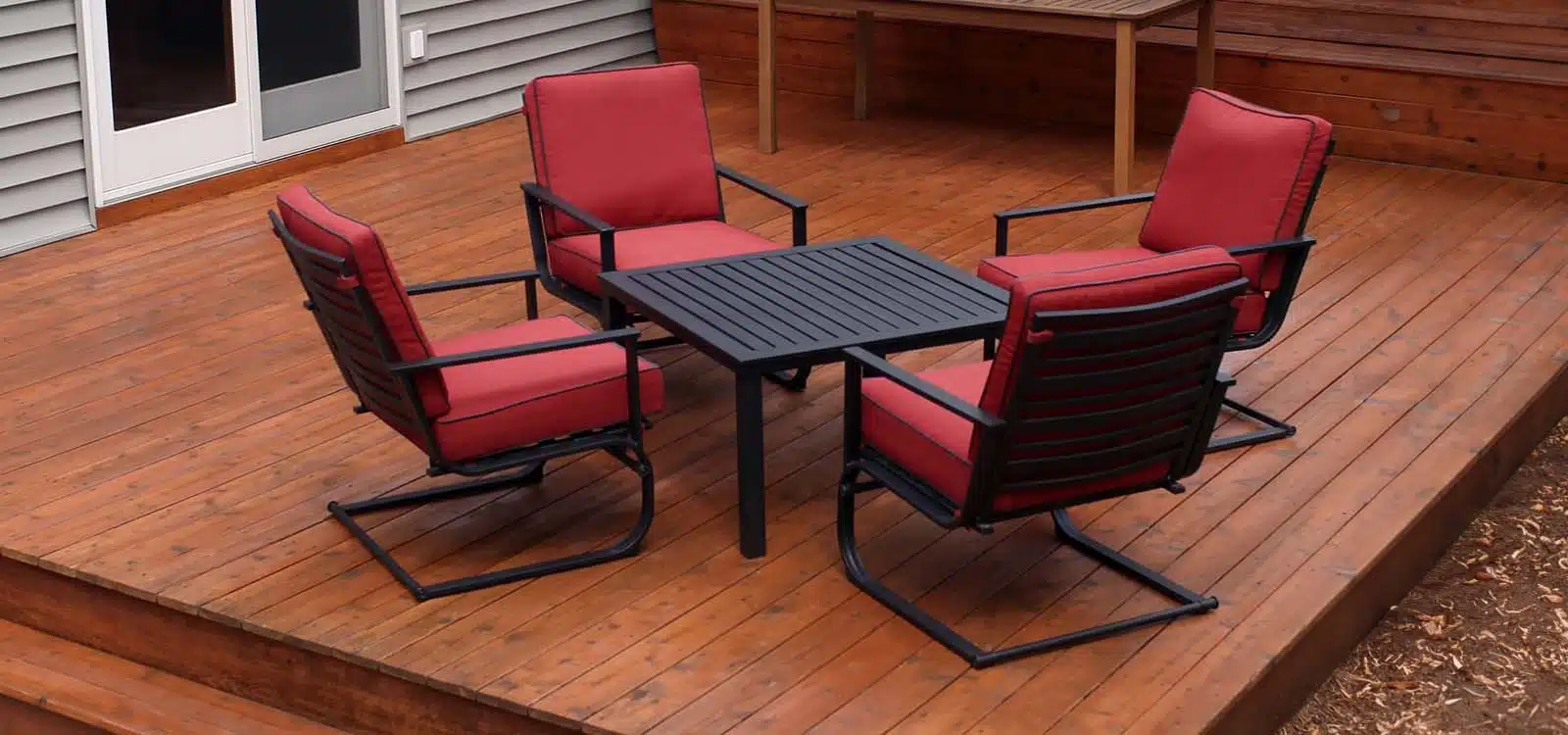 Four red cushioned patio chairs with black metal frames surround a square black metal table on a wooden deck, near a house with sliding doors.